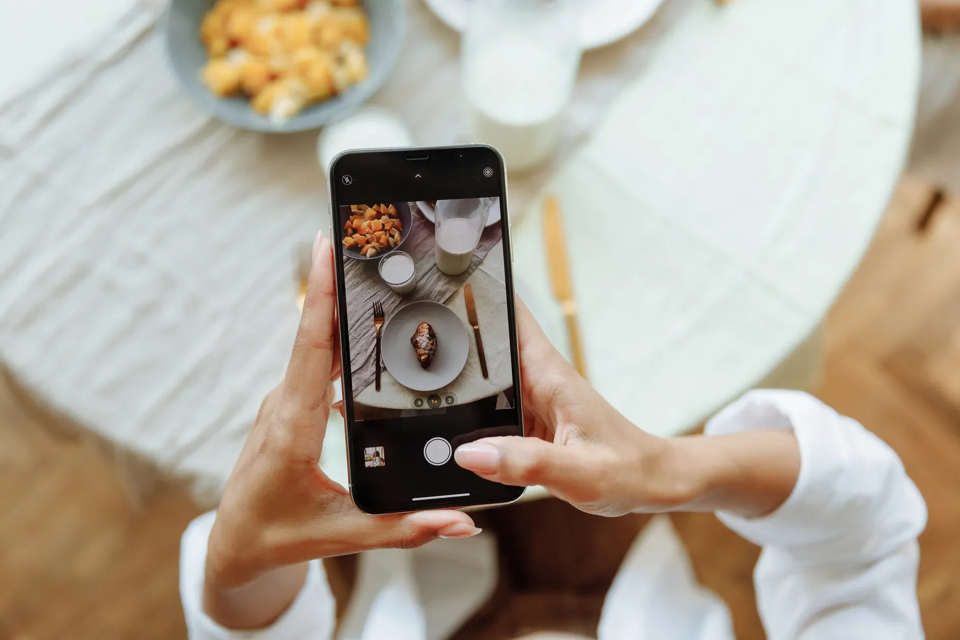 Eine Person fotografiert mit einem Smartphone Speisen und Geschirr auf einem hellen Tisch.